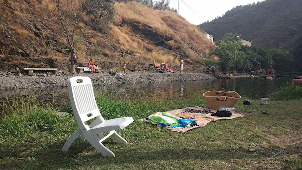 Silla blanca en la zona de césped junto a la piscina natural de Valero, con bañistas al fondo.