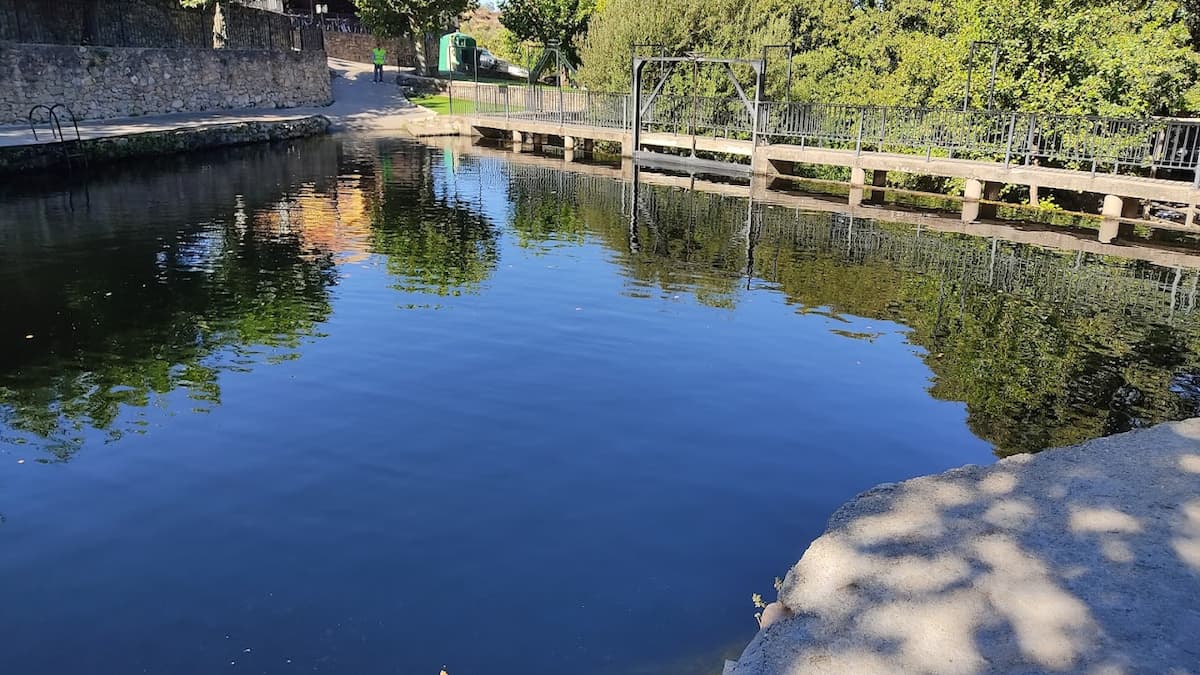 Reflejo del agua en la piscina natural de Casas del Monte con un puente peatonal de fondo.