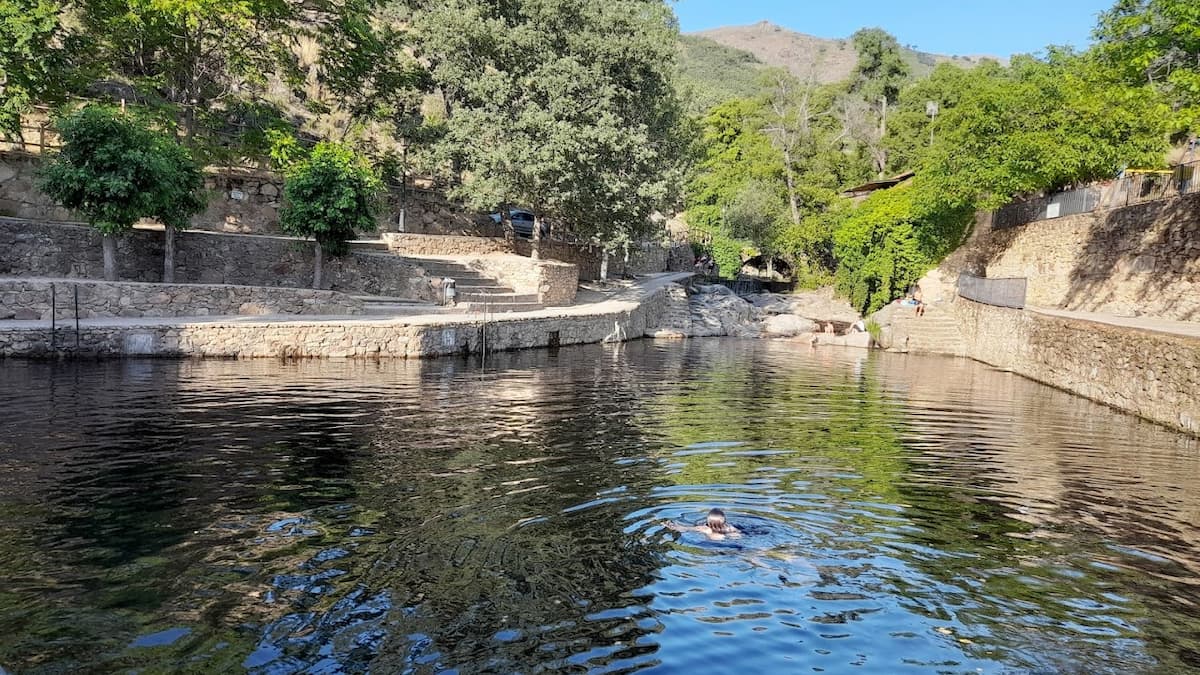 Piscina natural con agua cristalina, muros de piedra y visitantes disfrutando de un baño.