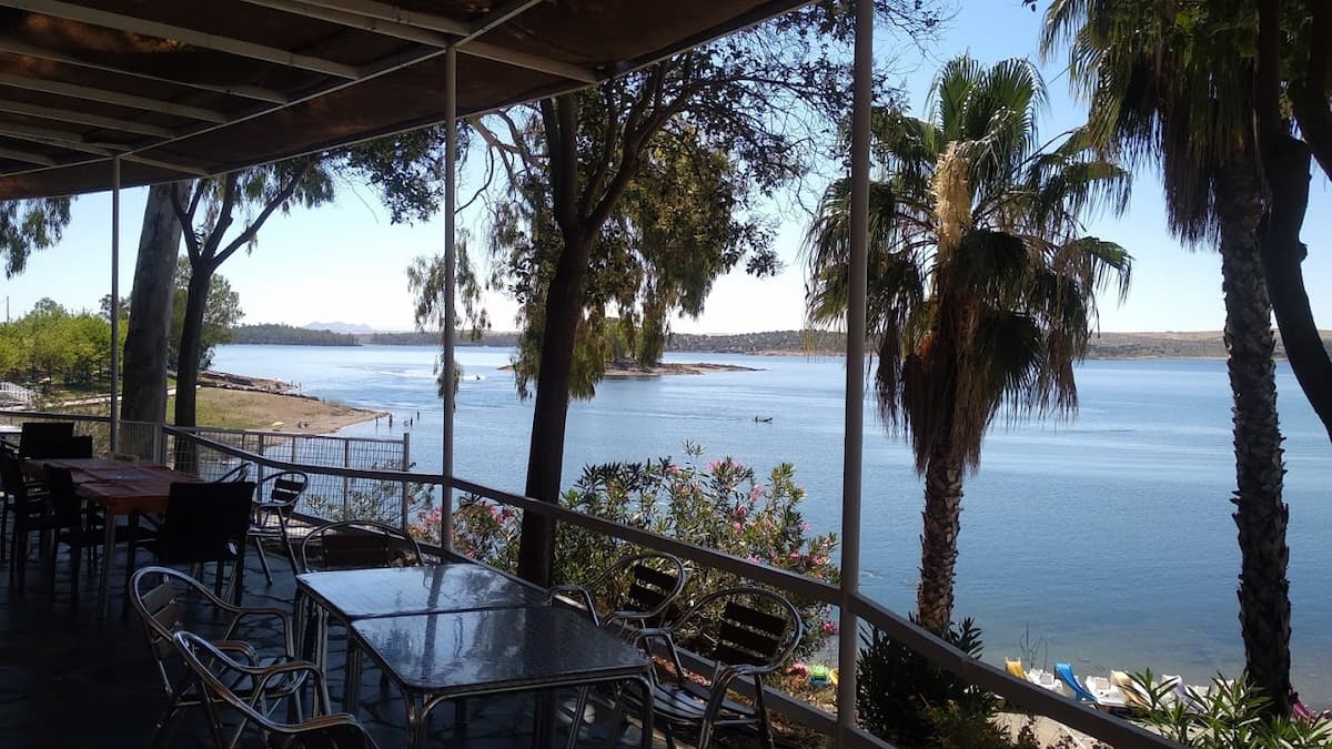 Terraza con mesas y sillas con vistas al embalse de Orellana, rodeada de árboles y vegetación.