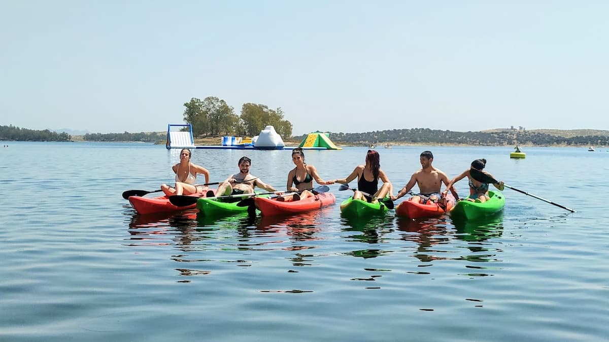 Grupo de jóvenes practicando kayak en el embalse de Orellana, con un parque acuático flotante en el fondo.
